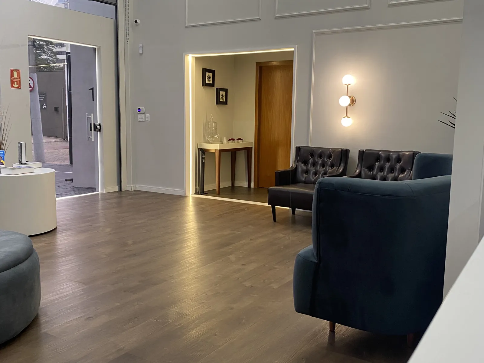 Sala de espera com luminárias em anel na parede, poltrona de veludo azul-petróleo e piso de madeira