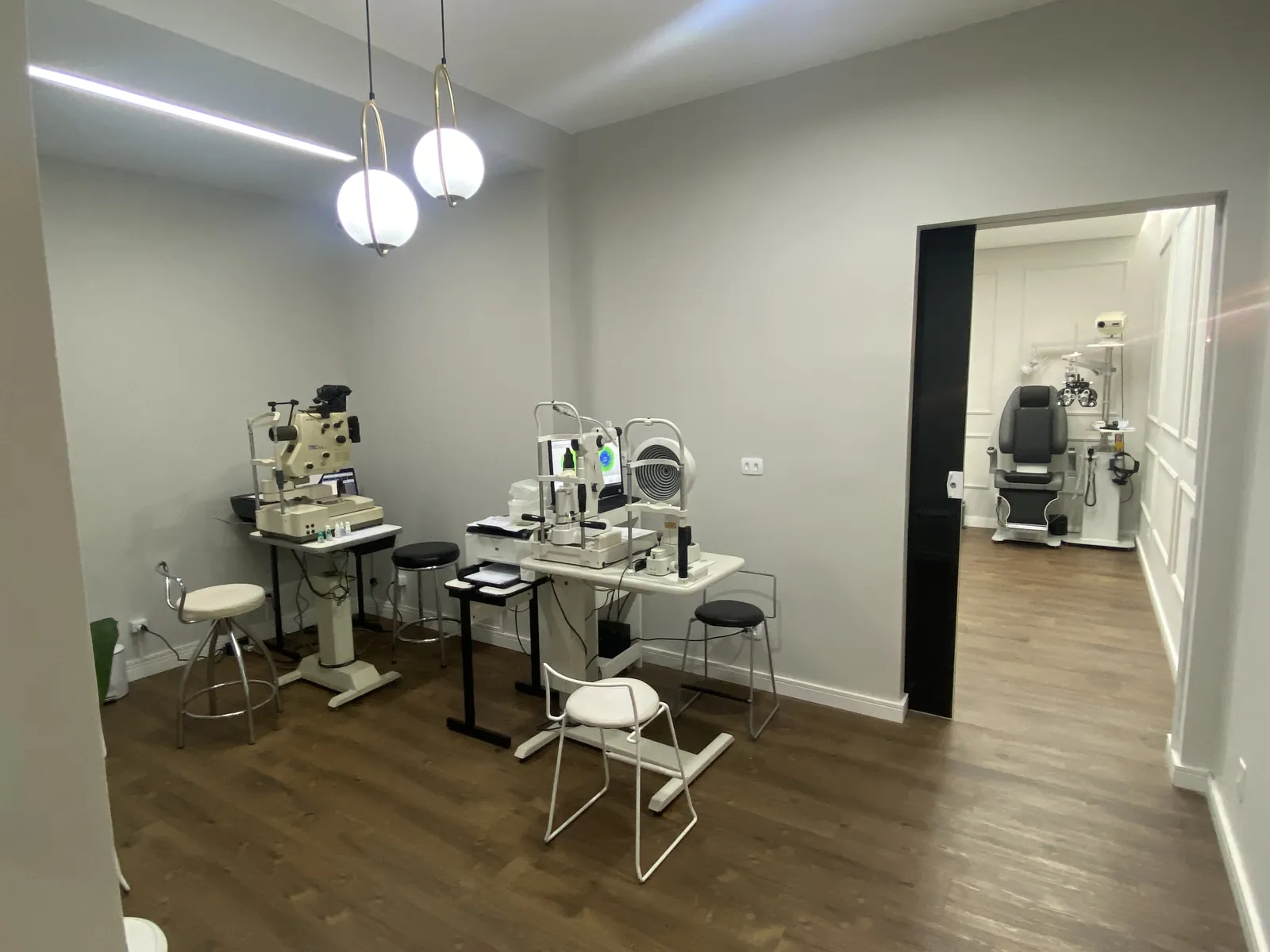Sala de exames com lâmpada de fenda, mesa branca, cadeiras e a porta aberta para o consultório